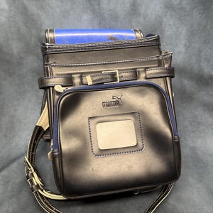 May include: A black Puma school backpack with a rectangular front pocket and a blue trim. The backpack has a buckle closure and adjustable straps. The Puma logo is visible on the front pocket.