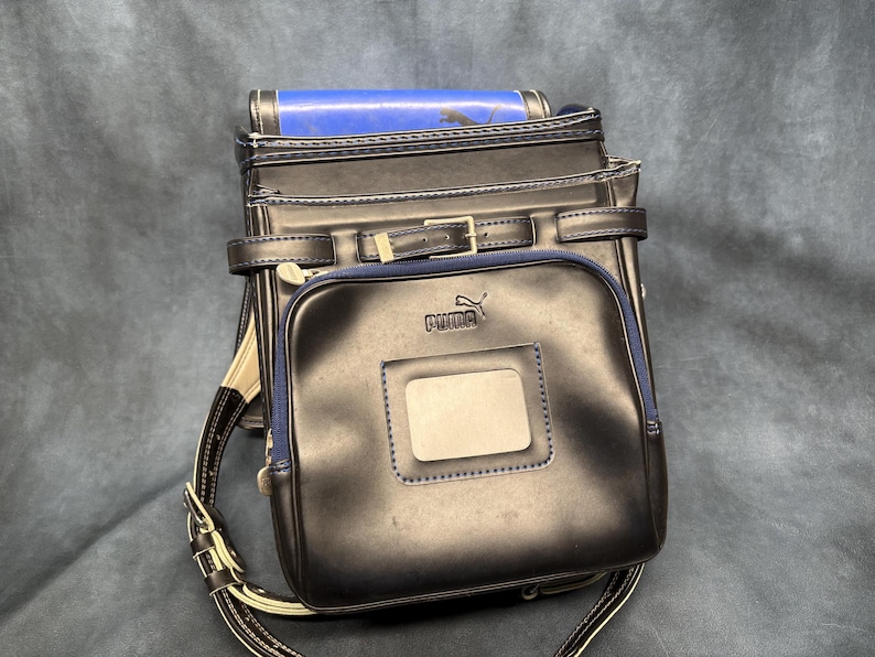May include: A black Puma school backpack with a rectangular front pocket and a blue trim. The backpack has a buckle closure and adjustable straps. The Puma logo is visible on the front pocket.