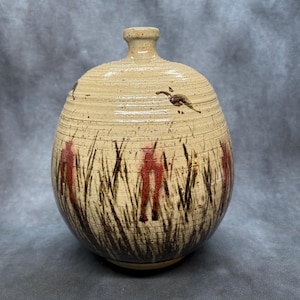 May include: A beige ceramic vase with a rounded body and a narrow neck. The vase is decorated with a design of tall, brown and red grass and flying birds. The vase has a textured surface.