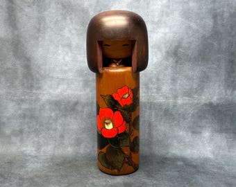 Puede incluir: Muñeca Kokeshi japonesa de madera con cabeza marrón y cuerpo cilíndrico. El cuerpo está pintado con flores de camelia rojas y hojas verdes sobre un fondo marrón. La muñeca tiene un diseño sencillo y minimalista.