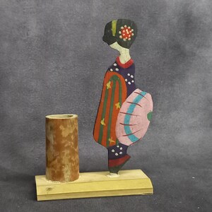 May include: A wooden figurine of a woman in a colorful kimono holding a pink and blue umbrella. The figurine is standing on a wooden base with a brown cylindrical container.