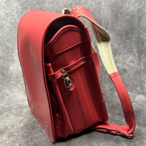 May include: A red leather school backpack with a white strap and a silver buckle. The backpack has a flap with a zipper and a pocket on the front.