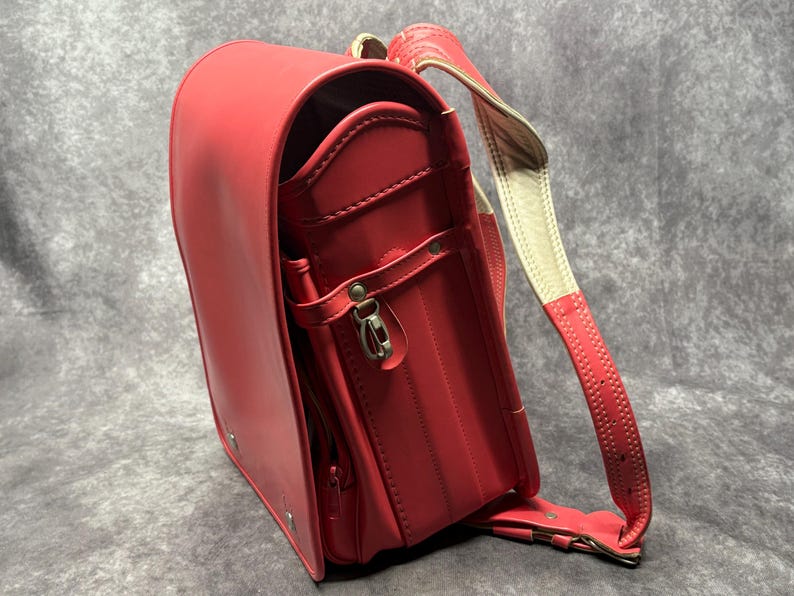 May include: A red leather school backpack with a white strap and a silver buckle. The backpack has a flap with a zipper and a pocket on the front.