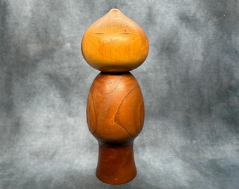 Könnte beinhalten: Eine Kokeshi-Puppe aus Holz mit rundem Kopf und zylindrischem Körper. Der Kopf ist heller braun als der Körper, mit zwei kleinen, horizontalen Augenschlitzen. Der Körper hat eine glatte, polierte Oberfläche, und die Puppe ist etwa 18 cm hoch.