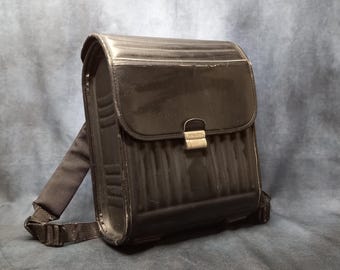 May include: Black backpack with a rectangular shape and a flap closure. The bag has a silver-colored clasp and adjustable shoulder straps. The backpack is made of a shiny, possibly leather-like material.