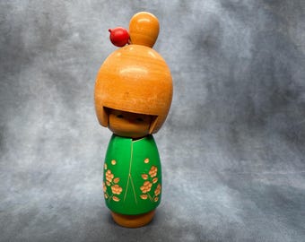 Puede incluir: Muñeca Kokeshi japonesa de madera con cabeza marrón claro y cuerpo verde. La cabeza tiene una pequeña bola roja encima. El cuerpo verde está decorado con motivos florales naranjas. La muñeca está de pie.
