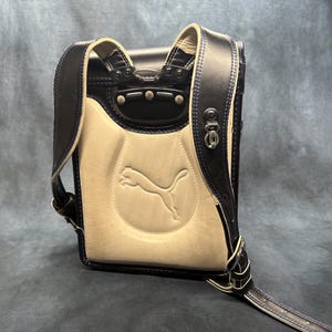 May include: A navy blue and cream-colored backpack with the Puma logo embossed on the front. The backpack features navy blue straps and trim with silver hardware. The stitching is navy blue.