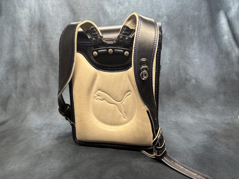 May include: A navy blue and cream-colored backpack with the Puma logo embossed on the front. The backpack features navy blue straps and trim with silver hardware. The stitching is navy blue.
