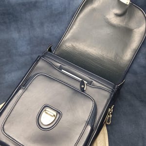 May include: A black leather satchel bag with a flap closure and a silver buckle. The bag has a zippered pocket on the front and a small pocket on the inside.