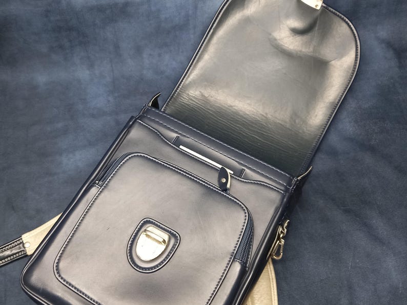 May include: A black leather satchel bag with a flap closure and a silver buckle. The bag has a zippered pocket on the front and a small pocket on the inside.