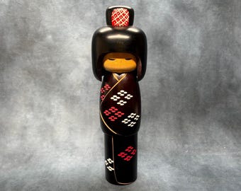 Puede incluir: Una muñeca Kokeshi japonesa con acabado lacado negro. La muñeca presenta una cabeza redondeada con un diseño rojo y blanco en la parte superior, una cara de madera y un kimono oscuro adornado con diseños florales blancos y rojos. La muñeca mide unos 25 cm de altura.