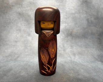 May include: A wooden Kokeshi doll with a dark brown finish. The doll features a rounded head and a cylindrical body. The face is painted with simple features. The body is decorated with etched wheat stalks in a lighter brown color.