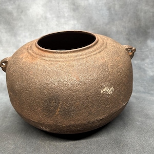 May include: A round, dark brown cast iron pot with a textured, weathered surface. The vessel features a wide, open circular top with a raised rim and two small, decorative side handles. The exterior shows signs of oxidation and age-related wear.