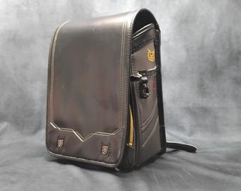 May include: Black leather school backpack with gold stitching and accents. The bag has a structured design with a flap closure, a side zipper, and decorative metal details. The interior is visible with a yellow zipper.