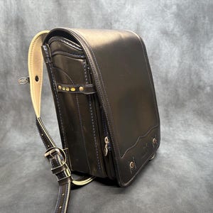 May include: Black leather school backpack with a rectangular shape and a curved top flap. The bag features a beige shoulder strap and gold-colored hardware. The design includes decorative stitching and a small front pocket.