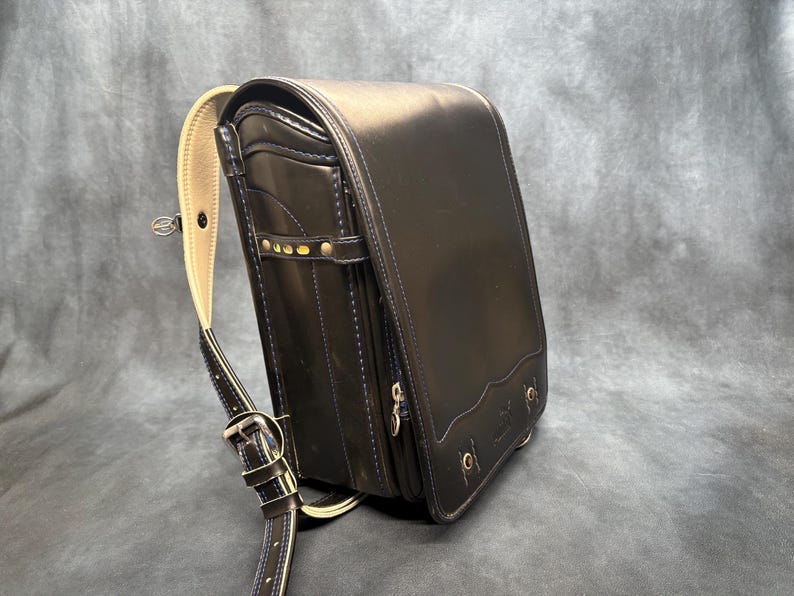 May include: Black leather school backpack with a rectangular shape and a curved top flap. The bag features a beige shoulder strap and gold-colored hardware. The design includes decorative stitching and a small front pocket.