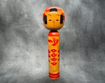 May include: A wooden Kokeshi doll with a round head and cylindrical body. The head is painted with a red top, black hair, and a serene face. The body is yellow with red floral and script designs.