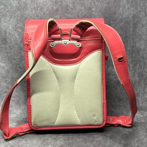 May include: A pink and white leather school backpack with adjustable straps. The backpack has a flap with a silver buckle and a white leather front panel.