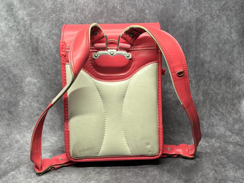 May include: A pink and white leather school backpack with adjustable straps. The backpack has a flap with a silver buckle and a white leather front panel.