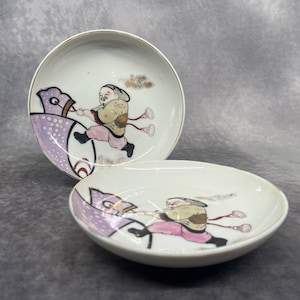 Könnte beinhalten: Zwei weiße Keramikteller mit einem lila und rosa Design. Jeder Teller zeigt eine handgemalte Illustration einer Person, die ein mythisches Wesen reitet.