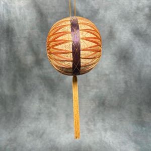 Puede incluir: Adorno esférico hecho a mano con un diseño complejo. El adorno presenta una base de hilo beige, superpuesta con hilo naranja y morado en un patrón geométrico. Una larga borla dorada cuelga de la parte inferior, y una cuerda dorada está en la parte superior.