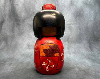 Puede incluir: Muñeca Kokeshi japonesa de madera con cabeza negra y cuerpo rojo. El cuerpo de la muñeca está adornado con motivos florales blancos y un diseño tallado. La cabeza de la muñeca tiene una pequeña gorra roja.