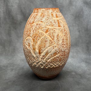 Puede incluir: Un jarrón de cerámica hecho a mano con una superficie moteada y texturizada. El jarrón presenta una paleta de colores crema y naranja con un diseño de hojas detallado. El jarrón tiene forma ovalada y es adecuado para la decoración del hogar.