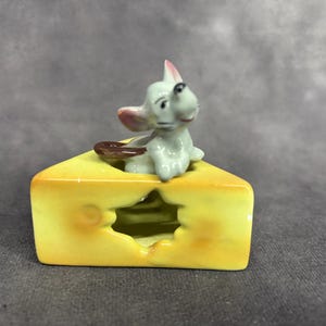 May include: A ceramic figurine of a white mouse sitting on a yellow wedge of cheese. The mouse has pink ears and a brown tail.