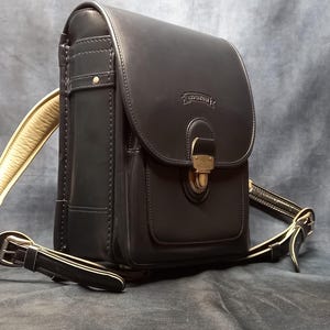 May include: A black leather satchel with a gold buckle and a strap. The bag has a flap with a gold buckle and a small pocket on the front. The bag has a long strap that can be adjusted to be worn over the shoulder or across the body.