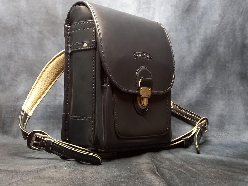 May include: A black leather satchel with a gold buckle and a strap. The bag has a flap with a gold buckle and a small pocket on the front. The bag has a long strap that can be adjusted to be worn over the shoulder or across the body.