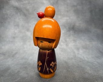 May include: Wooden Kokeshi doll with a rounded orange head and a dark brown body. The doll has a small red accent on top of its head and floral designs on the body. The doll's face is carved with simple features.