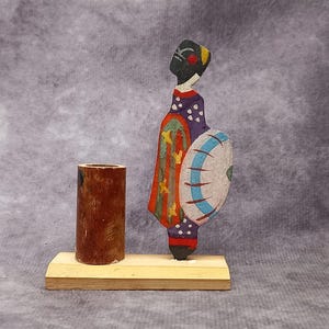 May include: A wooden stand with a brown cylindrical holder and a colourful painted figure of a person holding a shield. The figure is painted in shades of black, purple, green, orange, blue, and white.