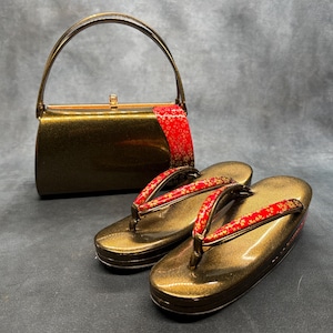May include: A gold-toned handbag with a matching pair of geta sandals. The sandals and bag feature red fabric accents with a floral pattern. The bag has a curved handle and a rectangular shape. The geta sandals are traditional Japanese footwear.