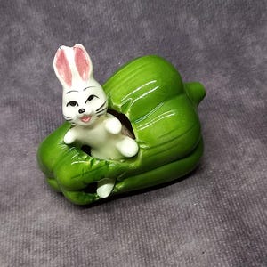 Może przedstawiać: Ceramiczna figurka białego królika wystająca z zielonego ceramicznego pieprzu. Królik ma różowe uszy i czarne oczy.
