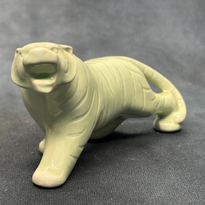 May include: A pale green ceramic tiger figurine in a prowling stance with its mouth open. The sculpture features a smooth, glossy finish and molded stripes across its body, capturing the animal in a dynamic, forward-moving position.