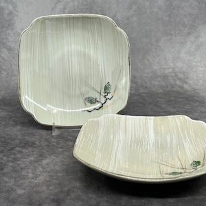 Puede incluir: Dos platos cuadrados de cerámica con fondo blanco y un sutil patrón de veta de madera. Los platos tienen un borde elevado y un delicado diseño floral en negro y verde.