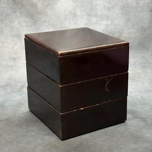 May include: A dark brown, three-tiered wooden box with a square shape. The box has a glossy finish and a lid that fits snugly on top. The stacked design suggests storage or organizational use.