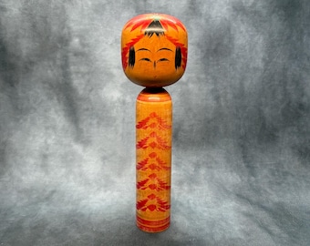 May include: A wooden Japanese Kokeshi doll with a round head and cylindrical body. The doll features a painted orange face with closed eyes and black hair. The body is orange with a red tree-like pattern. The doll is approximately 10 inches tall.