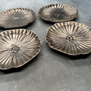 May include: Four silver-coloured, flower-shaped coasters. Each coaster features a central floral design and radiating lines. The coasters are arranged on a dark grey surface, highlighting their intricate details and metallic sheen.