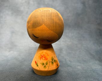 May include: Wooden Kokeshi doll with a round head and a conical body. The doll's face features closed eyes and long eyelashes. The body is decorated with a painted rose design in shades of orange and green. The doll is made of light-colored wood.
