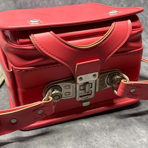 May include: A bright pink leather school bag with a metal clasp and adjustable straps. The bag has a vintage look and is in good condition.