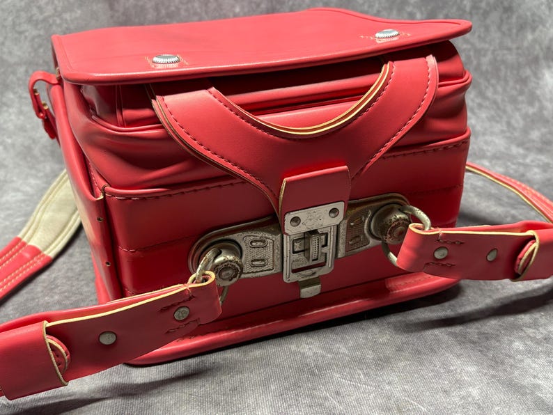 May include: A bright pink leather school bag with a metal clasp and adjustable straps. The bag has a vintage look and is in good condition.