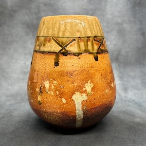 May include: A handmade ceramic vase with a unique, organic shape. The vase features a warm, earthy color palette with shades of brown, tan, and cream. Decorative cross-like patterns and drips of glaze add visual interest. A rustic, artisanal piece.