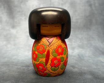 Może przedstawiać: Vintage japońska lalka Kokeshi z czarną, zaokrągloną głową i malowanym korpusem. Korpus lalki zdobi kwiatowy wzór w kolorze czerwonym i zielonym na żółtym tle. Lalka ma prosty, tradycyjny design.