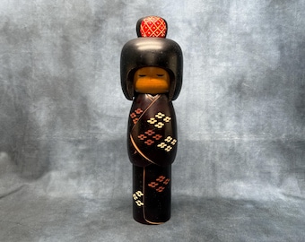 Puede incluir: Muñeca Kokeshi japonesa de madera con cabeza y cuerpo negros. La muñeca tiene una parte superior roja estampada y un diseño tipo kimono con patrones florales blancos y naranjas. La cara es de color naranja claro.