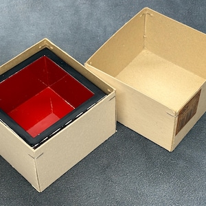 May include: Two-piece box set. The outer box is made of light brown cardboard. The inner box is red with a black border. The lid is off, revealing the interior. The boxes are on a gray surface.