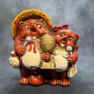 Peut inclure: Une figurine en céramique représentant deux chiens viverrins tanuki au pelage brun et au ventre crème. Le tanuki de gauche porte un chapeau de paille jaune et tient un micro argenté. Celui de droite a un nœud rouge et tient un trophée doré.