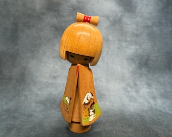 May include: A wooden Kokeshi doll with a smooth, light brown finish, featuring a bobbed hairstyle and a small red bow with white dots on top. The doll wears a kimono painted with a mountain landscape scene in green, white, and brown tones.