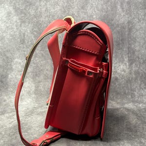 May include: A red leather school backpack with gold accents and a white trim. The backpack has a large main compartment and several smaller pockets.