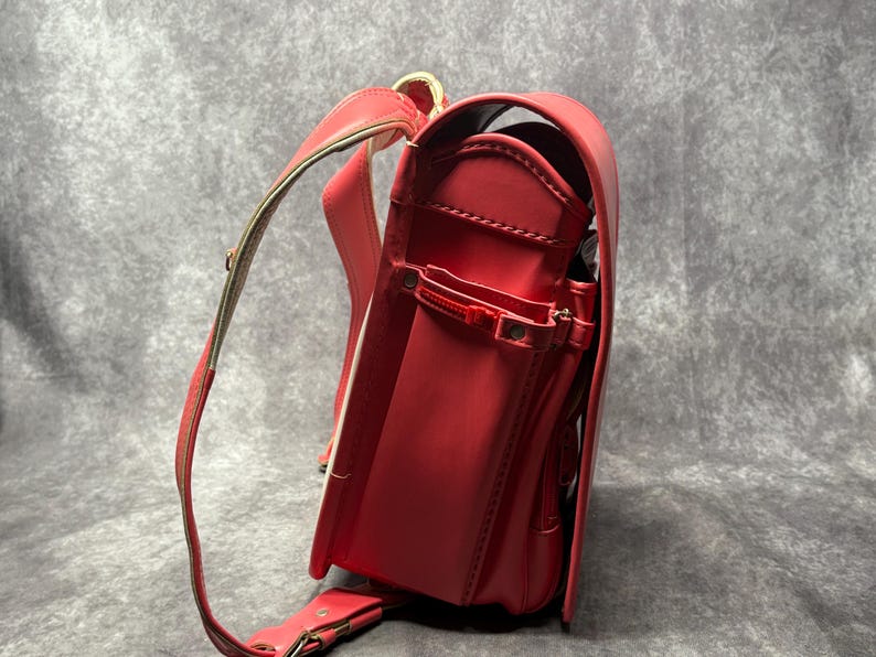 May include: A red leather school backpack with gold accents and a white trim. The backpack has a large main compartment and several smaller pockets.